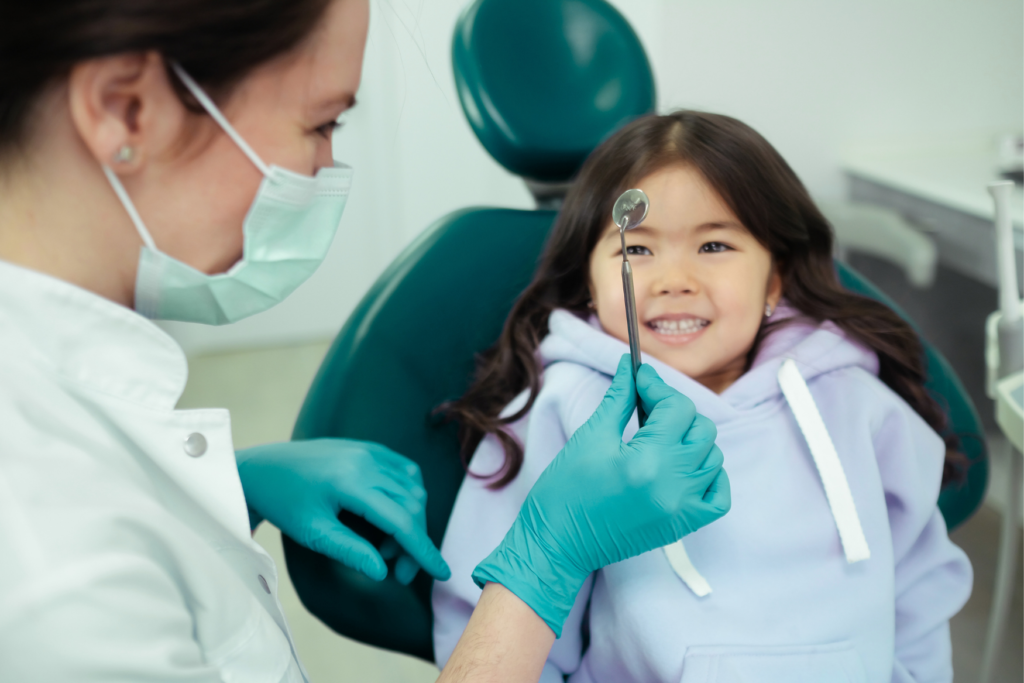A dentist showing her dental tool to a smiling young girl who is seated in a dental chair